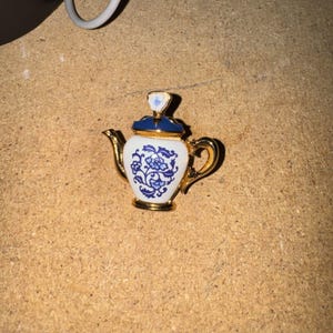 May include: A gold-toned metal brooch shaped like a miniature teapot. The body is white with a blue floral pattern, featuring a dark blue lid with a small white finial. The handle, spout, and base are polished gold-tone metal, creating a detailed accessory.