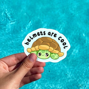 May include: A sticker depicting a cartoon turtle with a brown shell and green face and limbs. The sticker has the text "helmets are cool" in white font on a blue background. The sticker is held against a blue water background.