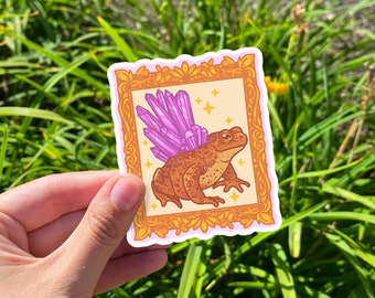 Puede incluir: Una pegatina con un sapo de dibujos animados con alas de cristal morado en un marco dorado. El sapo es marrón con la barriga de color naranja claro y está sentado sobre un fondo naranja claro. La pegatina está sobre una superficie verde cubierta de hierba.