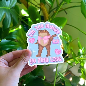 May include: A blue sticker with a cartoon frog wearing a pink dress and holding a pink purse. The text on the sticker reads "I'm too much? Go find less."