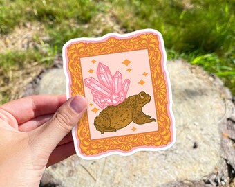 Puede incluir: Una pegatina con un sapo de dibujos animados con cristales rosas en la espalda, enmarcado en un marco dorado sobre un fondo rosa. La pegatina se sostiene en una mano sobre un fondo borroso de hierba verde y un tocón de árbol.