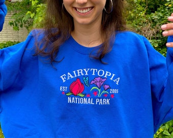 May include: A royal blue sweatshirt with a white embroidered design that says "Fairytopia Est. 2005 National Park" with colorful flowers and a butterfly.