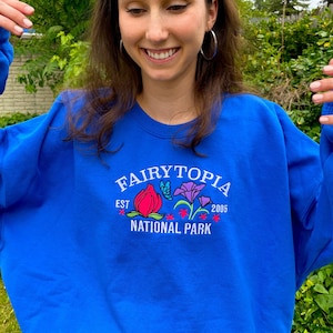 May include: A royal blue sweatshirt with a white embroidered design that says "Fairytopia Est. 2005 National Park" with colorful flowers and a butterfly.