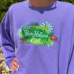 May include: A lilac sweatshirt featuring the embroidered phrase "Pixie Hollow Girl" in a green oval, framed by white daisies and red flowers. The long-sleeved top is made of a soft material.