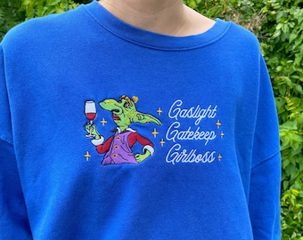 May include: A royal blue sweatshirt featuring a green cartoon character holding a glass of wine. The text "Gaslight Gatekeep Girlboss" is embroidered in white on the sweatshirt.
