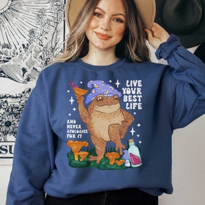 May include: A navy blue sweatshirt with a cartoon frog wearing a purple and blue hat and holding a glass of liquid. The frog is surrounded by mushrooms and the text "Live your best life" and "And never apologize for it" is printed on the sweatshirt.