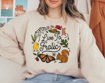 May include: A cream-colored sweatshirt with a whimsical design featuring a butterfly, a squirrel, a snail, a chick, and flowers. The text "I Live To Frolic" is written in a decorative font.