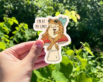 May include: A sticker featuring a cartoon frog with fairy wings, holding a glass of wine, and sitting on a mushroom. The text "I'M AT MY LIMIT" is above the frog. The sticker has a white border and is held against a green background.
