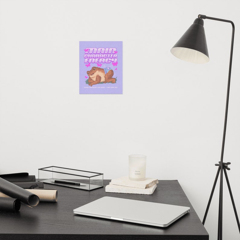 May include: A purple background with a cartoon illustration of a brown frog wearing a blue swimsuit. The text "Main Character Energy" is written in pink letters above the frog.