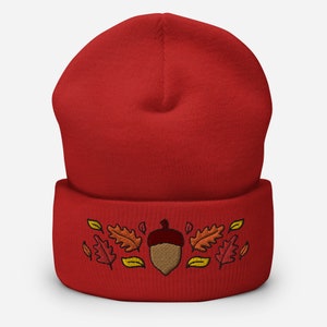 May include: A red beanie hat featuring an embroidered acorn and autumn leaves in brown, orange, and yellow. The hat is made of a soft, knitted material, suitable for autumn and winter wear.