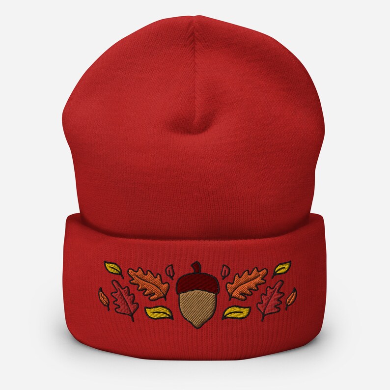 May include: A red beanie hat featuring an embroidered acorn and autumn leaves in brown, orange, and yellow. The hat is made of a soft, knitted material, suitable for autumn and winter wear.