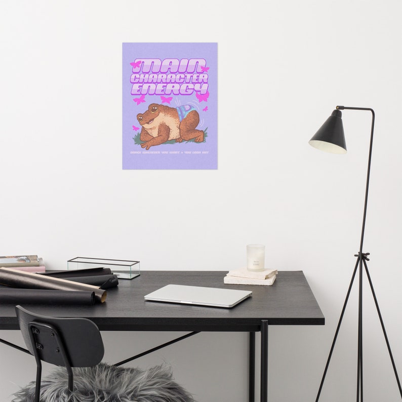 May include: A purple poster with the text "Main Character Energy" in pink letters. A cartoon frog with a blue butterfly on its back is sitting on a green patch of grass. Pink butterflies are flying around the frog.