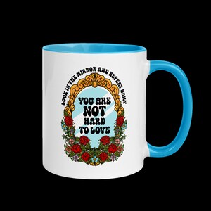 May include: White ceramic mug with a blue rim and handle. The mug features a colorful illustration of a mirror with the text "Look in the mirror and repeat daily. You are not hard to love."