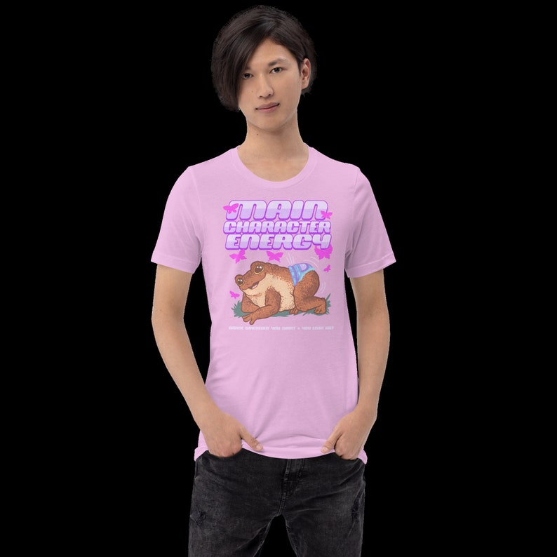 K&ouml;nnte beinhalten: Ein pinkfarbenes T-Shirt mit einer Cartoon-Froschfigur, die eine Windel tr&auml;gt, und dem Text "Main Character Energy" in pinker und violetter Schrift.