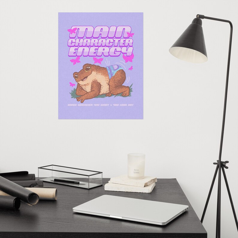 May include: Purple background with a cartoon frog wearing a nappy and pink butterflies. The text "MAIN CHARACTER ENERGY" is in a retro font. The text below the frog says "Embrace everything you always + you know what"