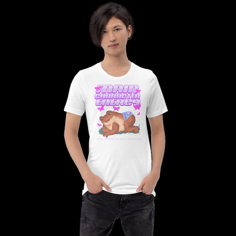 K&ouml;nnte beinhalten: Wei&szlig;es T-Shirt mit einem Grafikdruck einer Frosch, der eine Windel tr&auml;gt, und dem Text "Main Character Energy" in rosa Buchstaben.