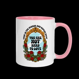 May include: White ceramic mug with a pink rim and handle. The mug features a graphic of a mirror with the text "Look in the mirror and repeat daily. You are not hard to love." The mirror is surrounded by a floral wreath.