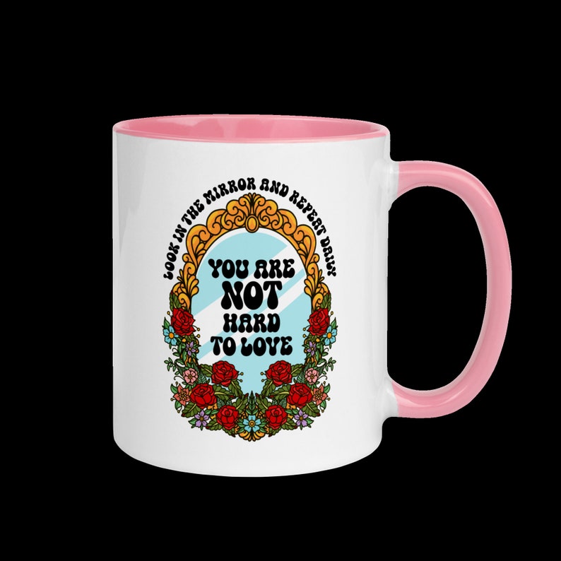 May include: White ceramic mug with a pink rim and handle. The mug features a graphic of a mirror with the text "Look in the mirror and repeat daily. You are not hard to love." The mirror is surrounded by a floral wreath.