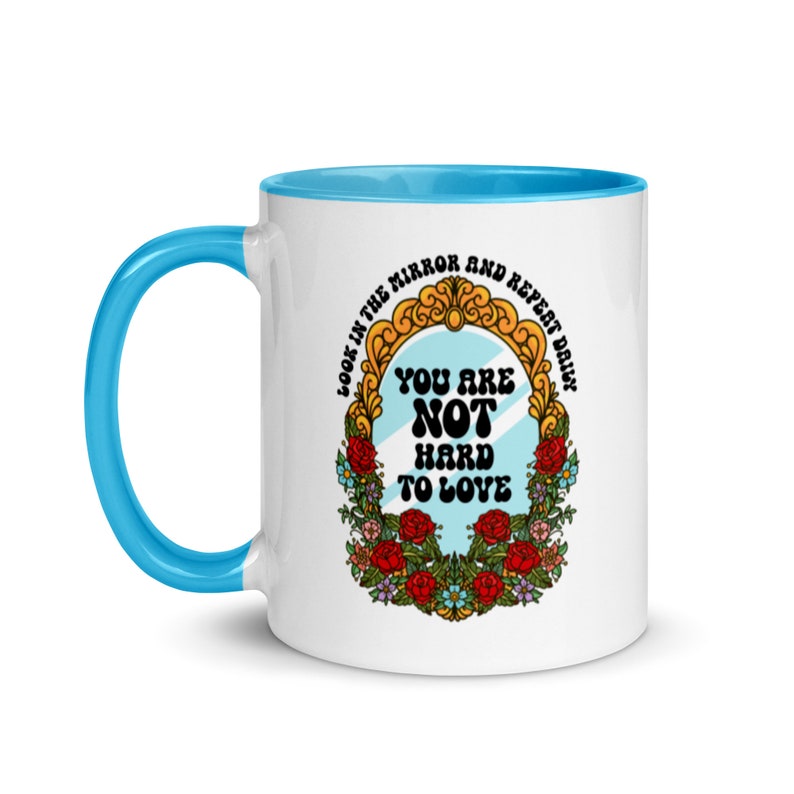 May include: White ceramic mug with a blue rim. The mug features a colorful illustration of a mirror with the text "Look in the mirror and repeat daily. You are not hard to love."