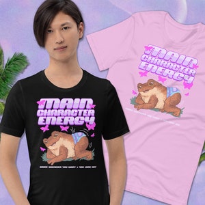 K&ouml;nnte beinhalten: Zwei T-Shirts mit einem Frosch, der eine Windel tr&auml;gt, und dem Text "Main Character Energy" in rosa Buchstaben mit Schmetterlingen drumherum. Die T-Shirts sind schwarz und rosa.