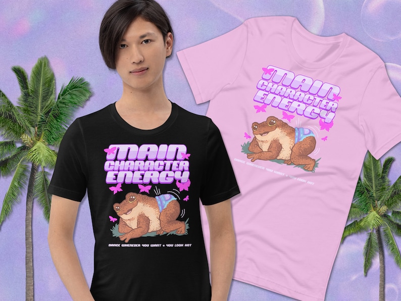K&ouml;nnte beinhalten: Zwei T-Shirts mit einem Frosch, der eine Windel tr&auml;gt, und dem Text "Main Character Energy" in rosa Buchstaben mit Schmetterlingen drumherum. Die T-Shirts sind schwarz und rosa.