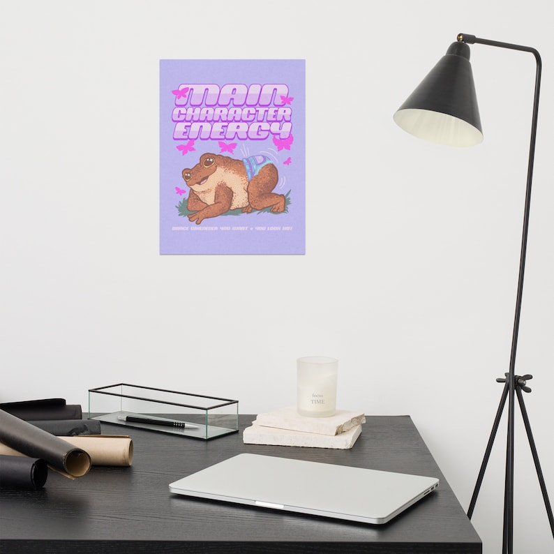 May include: A purple background with pink text that reads "Main Character Energy". A cartoon frog wearing a blue and white patterned outfit is sitting on a green leaf with pink butterflies flying around it.
