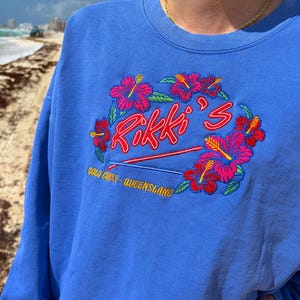 以下が含まれることがあります： 青色のスウェットシャツ。赤い筆記体で「Rikki's」と刺繍され、ピンクと赤のハイビスカスの花で囲まれています。「Gold Coast - Queensland」の文字も刺繍されています。カジュアルなスタイルです。