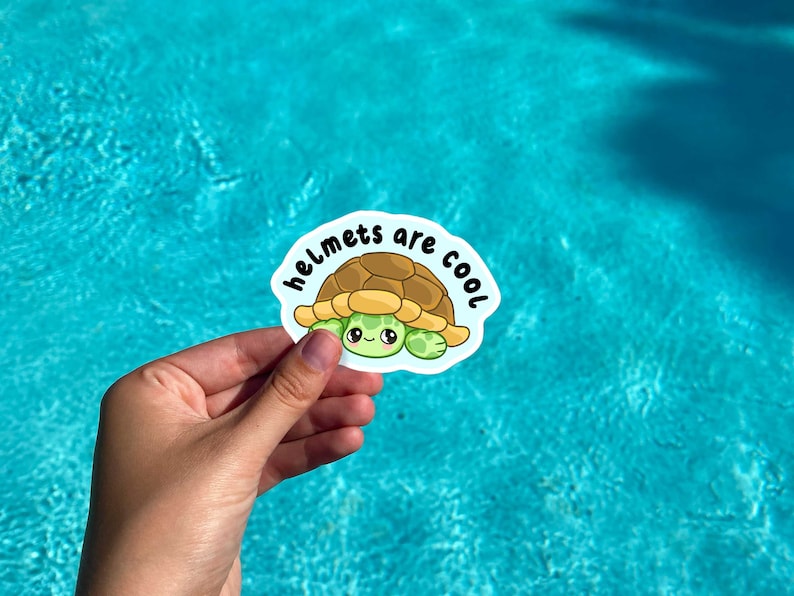 May include: A sticker depicting a cartoon turtle with a brown shell and green face and limbs. The sticker has the text "helmets are cool" in white font on a blue background. The sticker is held against a blue water background.