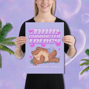 May include: A purple poster with the text "Main Character Energy" in pink letters. A cartoon frog wearing a blue and white patterned nappy is sitting on a green patch of grass. Pink butterflies are flying around the frog.