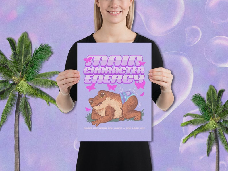 May include: A purple poster with the text "Main Character Energy" in pink letters. A cartoon frog wearing a blue and white patterned nappy is sitting on a green patch of grass. Pink butterflies are flying around the frog.