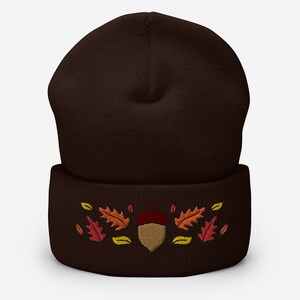 May include: A dark brown beanie hat featuring an embroidered acorn and autumn leaves in red, orange, and yellow. The hat is made of a soft, knitted material, suitable for the autumn and winter seasons.