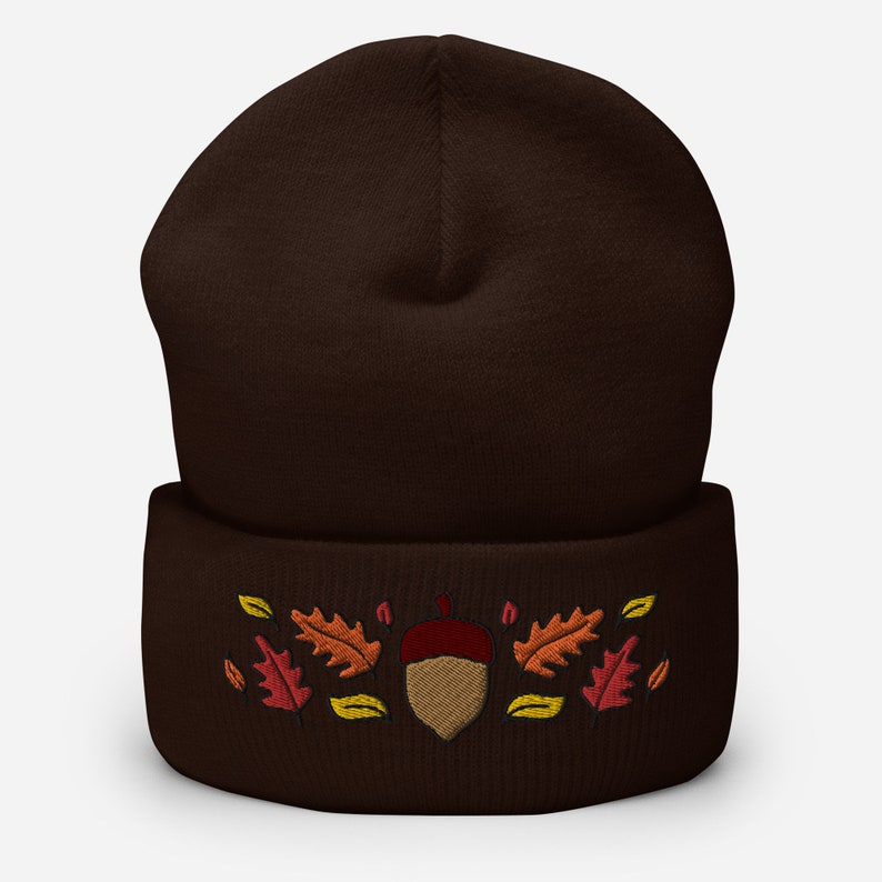 May include: A dark brown beanie hat featuring an embroidered acorn and autumn leaves in red, orange, and yellow. The hat is made of a soft, knitted material, suitable for the autumn and winter seasons.