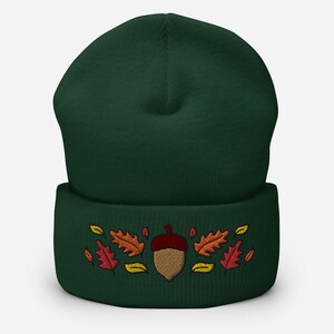 May include: A dark green beanie hat featuring an embroidered acorn and autumn leaves in red, orange, and yellow. The hat has a folded cuff, suitable for autumn and winter. The design evokes a seasonal theme.