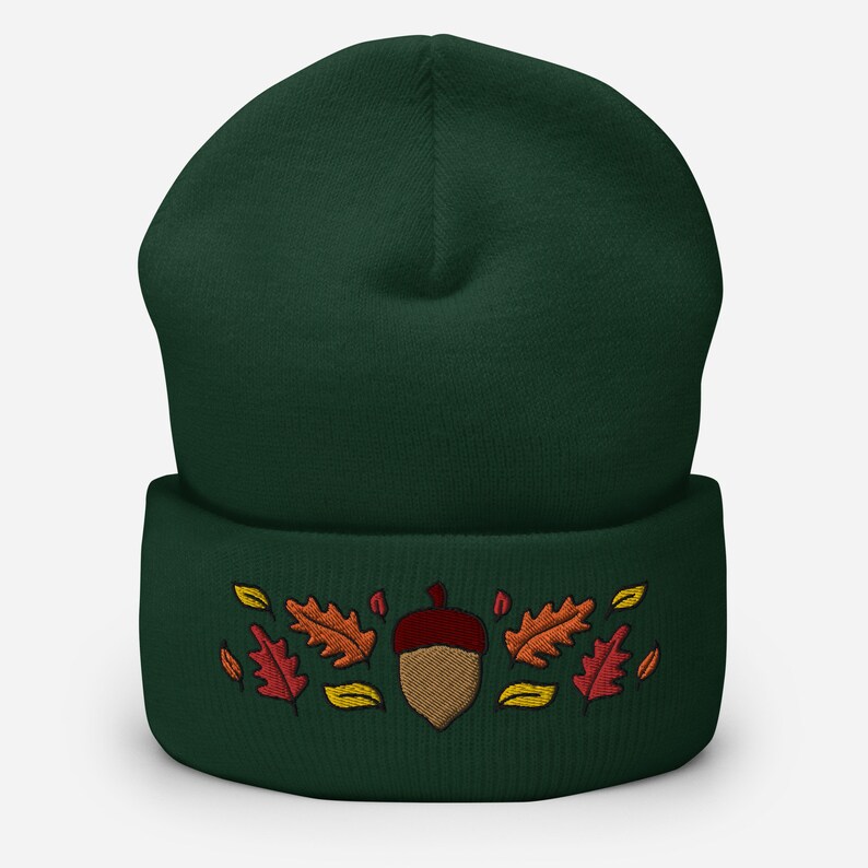 May include: A dark green beanie hat featuring an embroidered acorn and autumn leaves in red, orange, and yellow. The hat has a folded cuff, suitable for autumn and winter. The design evokes a seasonal theme.