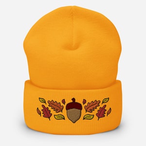 May include: Mustard yellow beanie hat featuring an embroidered acorn and autumn leaves in brown, orange, and yellow. A warm and comfortable accessory for the autumn season.