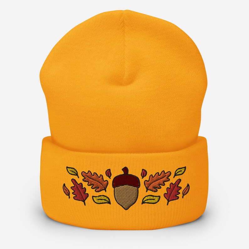 May include: Mustard yellow beanie hat featuring an embroidered acorn and autumn leaves in brown, orange, and yellow. A warm and comfortable accessory for the autumn season.