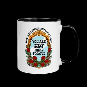 May include: White ceramic mug with a black rim and handle. The mug features a colorful illustration of a mirror with the text "Look in the mirror and repeat daily. You are not hard to love."