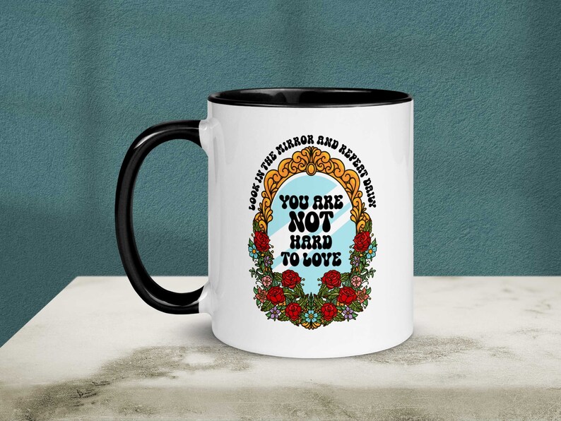 May include: White ceramic mug with a black rim and handle. The mug features a colorful illustration of a mirror with the text "Look in the mirror and repeat daily. You are not hard to love."