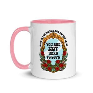 May include: White ceramic coffee mug with a pink rim. The mug features a colorful illustration of a mirror with the text "Look in the mirror and repeat daily. You are not hard to love."
