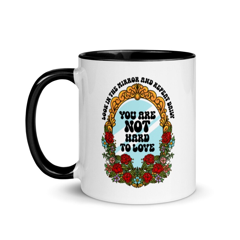 May include: White ceramic mug with a black rim and handle. The mug features a colorful illustration of a mirror with the text "Look in the mirror and repeat daily. You are not hard to love."