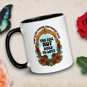 May include: White ceramic mug with a black handle. The mug has a colorful illustration of a mirror with the text "Look in the mirror and repeat daily: You are not hard to love."
