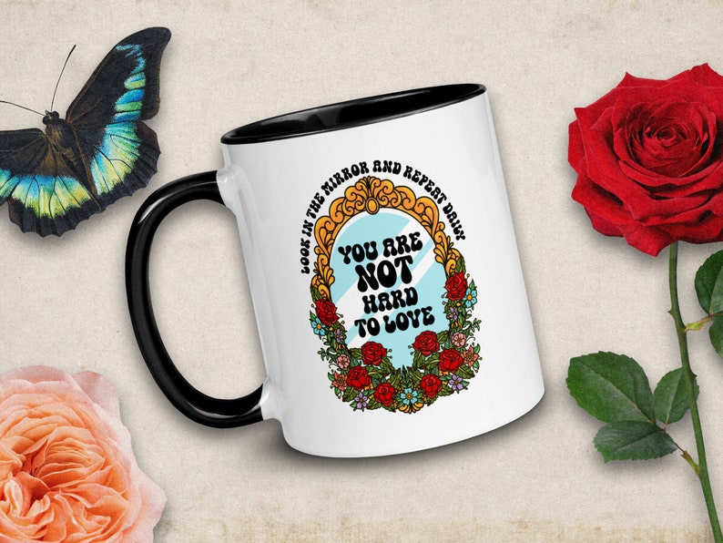 May include: White ceramic mug with a black handle. The mug has a colorful illustration of a mirror with the text "Look in the mirror and repeat daily: You are not hard to love."