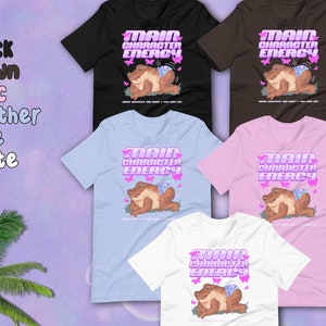 K&ouml;nnte beinhalten: Ein Set T-Shirts in verschiedenen Farben mit einem Cartoon-Frosch, der eine Windel tr&auml;gt und den Text "Main Character Energy". Der Frosch ist von rosa Schmetterlingen umgeben.