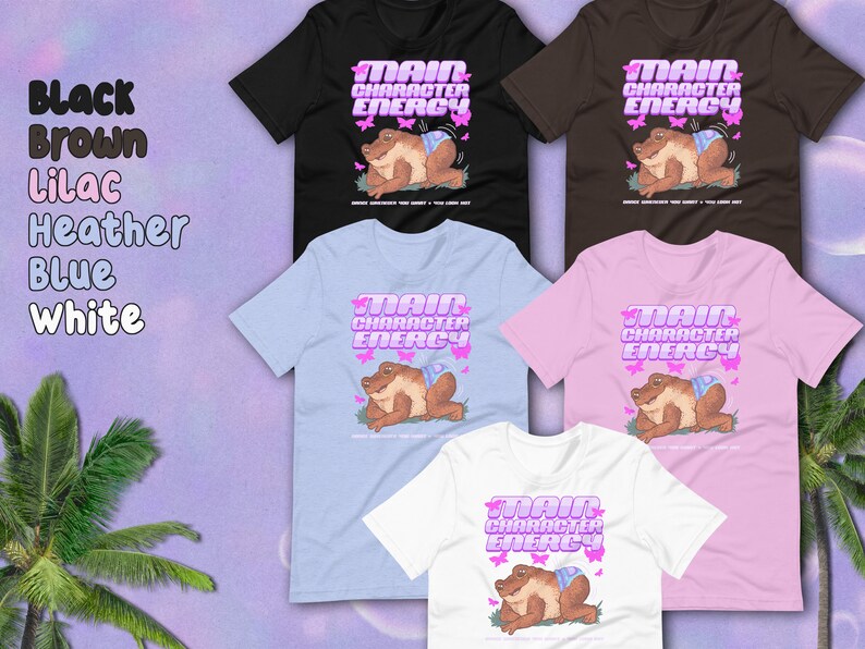 K&ouml;nnte beinhalten: Ein Set T-Shirts in verschiedenen Farben mit einem Cartoon-Frosch, der eine Windel tr&auml;gt und den Text "Main Character Energy". Der Frosch ist von rosa Schmetterlingen umgeben.