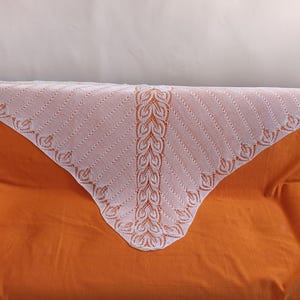 Puede incluir: Un chal triangular blanco con un delicado diseño calado. El chal presenta un patrón vertical central y un borde decorativo con motivos de hojas. Está colocado sobre un sofá naranja, creando un contraste de color y textura.