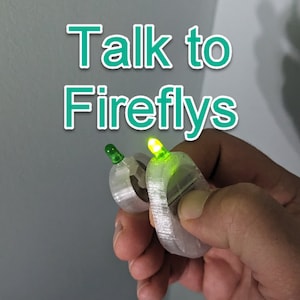 Könnte beinhalten: Zwei kleine, weiße, 3D-gedruckte Geräte mit grünen LED-Leuchten. Die Geräte sind ringförmig und haben einen Knopf an der Seite. Der Text "Talk to Fireflies" ist in grünen Buchstaben über den Geräten geschrieben.