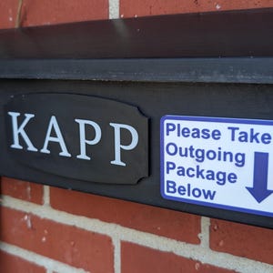 May include: Black mailbox with the name "KAPP" in white lettering. A sticker on the side reads "Please Take Outgoing Package Below" with a downward arrow. The mailbox is mounted on a red brick wall.