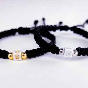 Peut inclure: Deux bracelets tressés noirs avec des perles blanches et des accents dorés et argentés. Les perles portent les lettres "S" et "M".