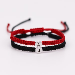 Pode incluir: Duas pulseiras de amizade com cord&otilde;es vermelhos e pretos, amarradas com um n&oacute;. As pulseiras t&ecirc;m pingentes de cora&ccedil;&atilde;o prateados com as letras "A" e "J" gravadas.