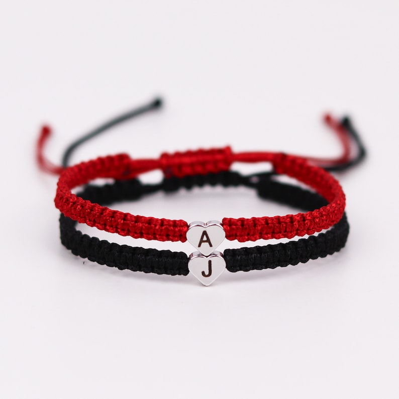 Pode incluir: Duas pulseiras de amizade com cord&otilde;es vermelhos e pretos, amarradas com um n&oacute;. As pulseiras t&ecirc;m pingentes de cora&ccedil;&atilde;o prateados com as letras "A" e "J" gravadas.